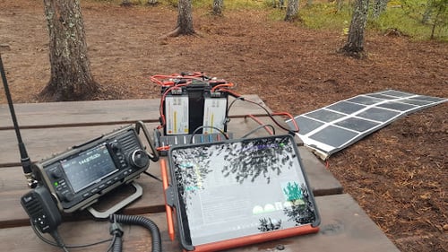 Lithium iron phosphate battery with two Genasun charge controllers mounted on the back with an Anderson Powerpole strip a tablet, ham radio on a wooden table with a 160W Crystalline Foldable Solar Panel charging the battery