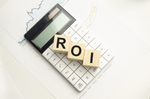 Wooden cubes spelling "ROI" placed on a calculator, with financial charts and graphs in the background, representing return on investment.