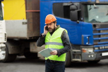 A worker in a safety vest and hard hat talks on the phone while holding documents, standing near trucks at a job site.