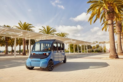 A bright blue GEM low-speed electric vehicle with PowerFilm solar panels installed on the roof is parked near a modern white surrounded by palm trees on a sunny day.