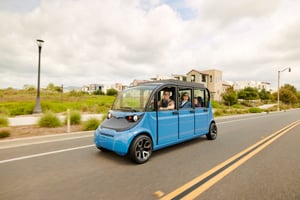 A blue GEM low-speed electric vehicle drives down a suburban street with several smiling passengers inside, passing modern houses and greenery under a cloudy sky.