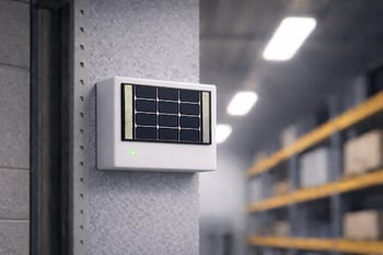 Wall-mounted indoor solar-powered device with a small front-facing panel and green indicator light, installed on a warehouse wall with shelving and overhead lights visible in the background.