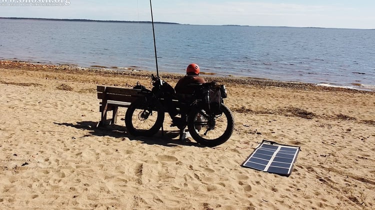 Portable crystalline solar panel deployed beside a bicycle-supported amateur radio station near a shoreline.